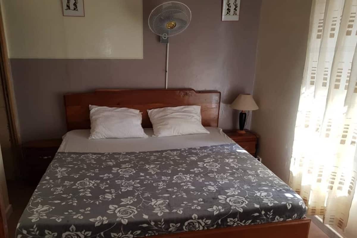A bed with a floral bedspread, a nightstand with a lamp, a wall-mounted fan, and a window with curtains.