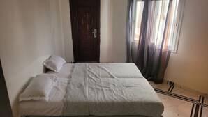 A bed with a patterned bedspread, two pillows, a wooden door, and a window with curtains. - Chez Medzo (Toubab Dialao)