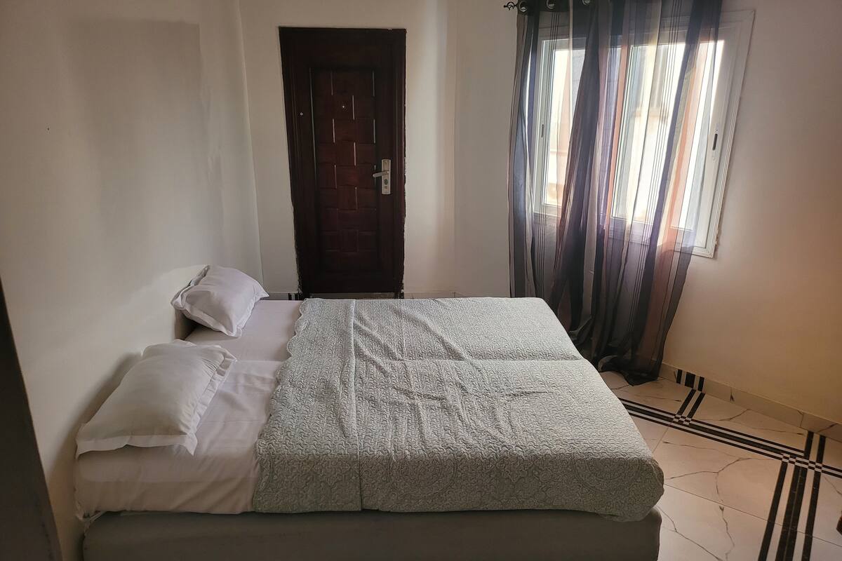 A bed with a patterned bedspread, two pillows, a wooden door, and a window with curtains.