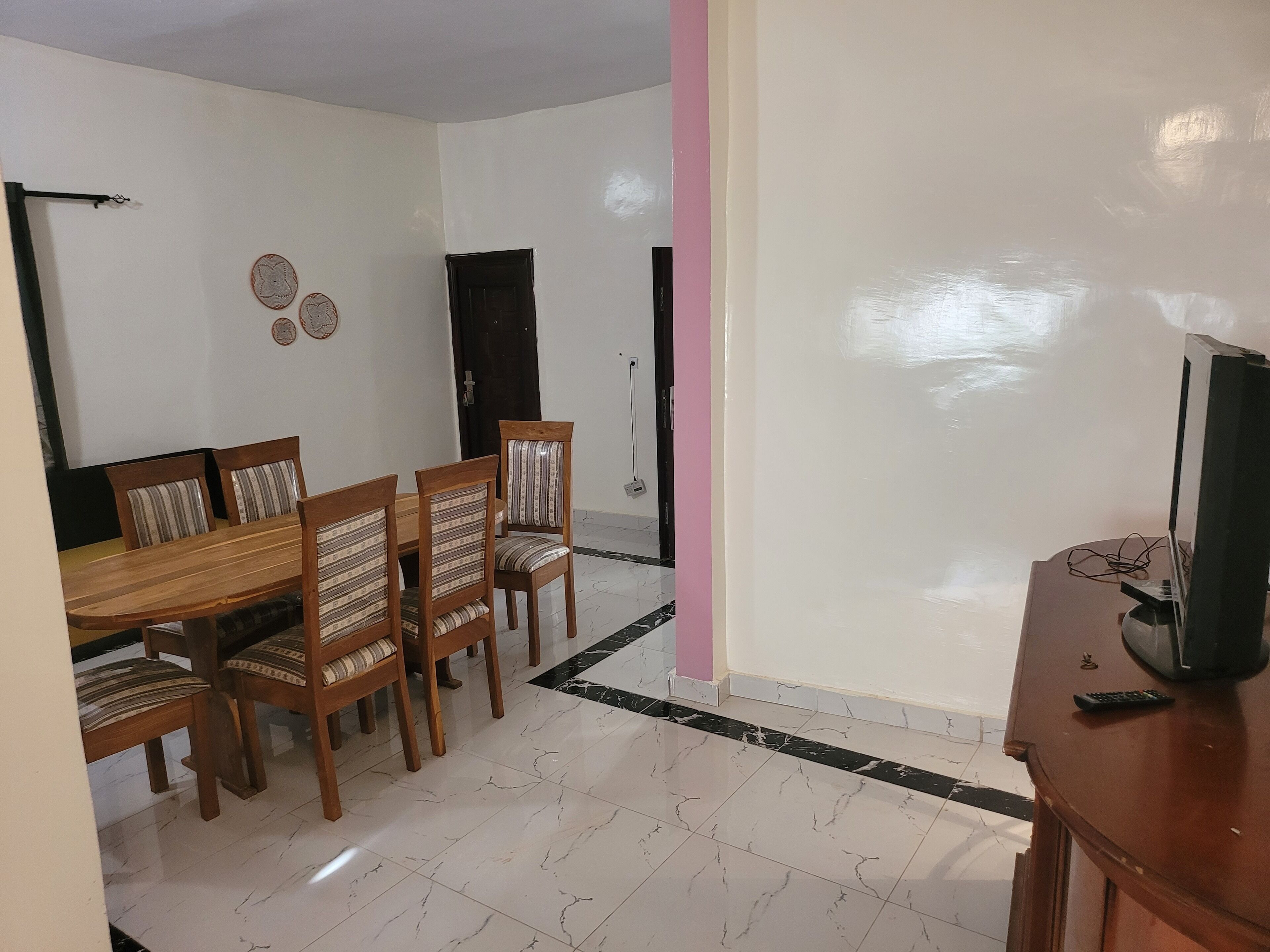 Un área de comedor con una mesa y sillas de madera, un televisor sobre un mueble bajo, y una pared de acento en color rosa.