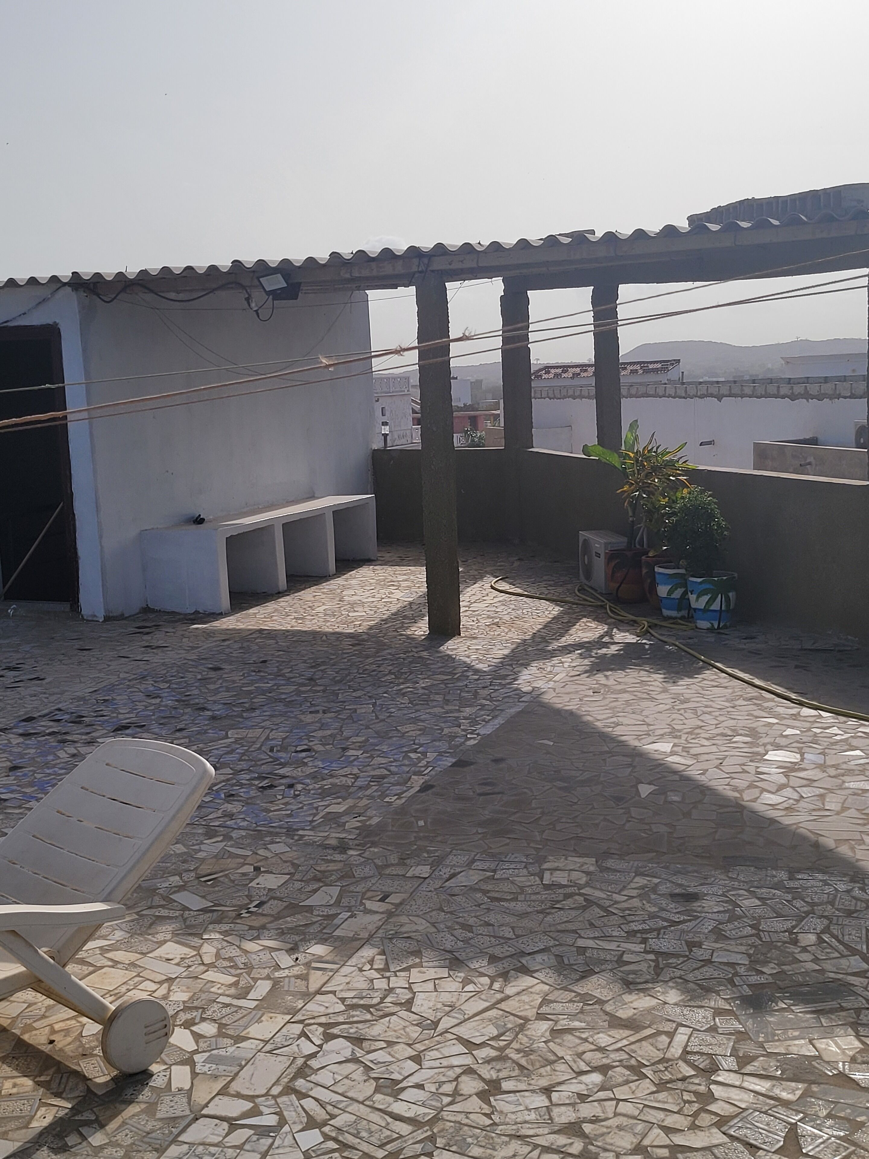 Una terraza en la azotea con suelo de baldosas, una silla blanca, plantas en macetas y una pérgola.