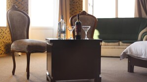 Una habitación con una mesita de noche de madera oscura que sostiene una botella, un vaso y una botella de agua. Se ve una silla y un sofá verde al fondo.