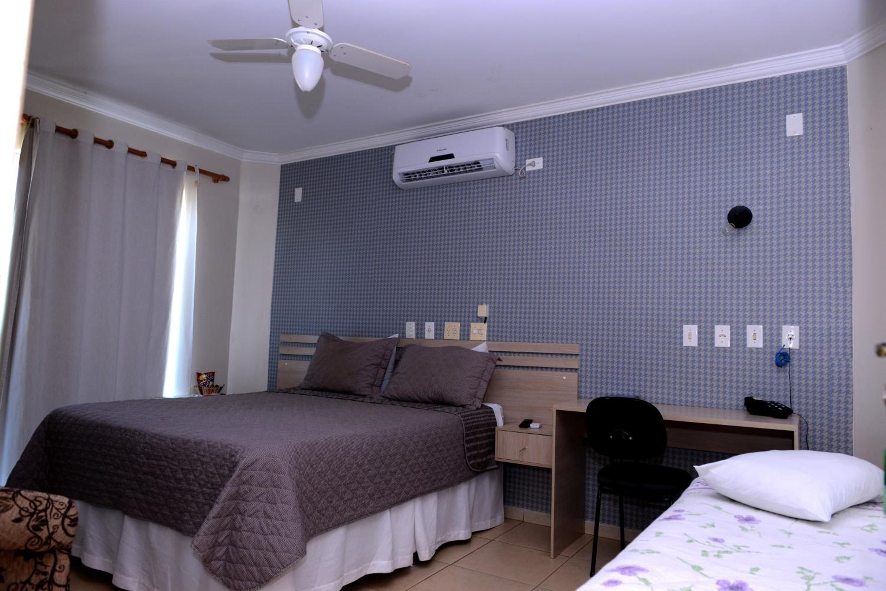 A bedroom with a bed, a desk, a chair, an air conditioning unit, and a window with curtains.