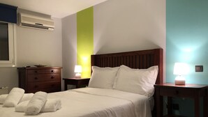 Una cama bien hecha con sábanas y almohadas blancas, una mesita de noche con lámpara, una mesita auxiliar de madera y un aire acondicionado instalado en la pared.