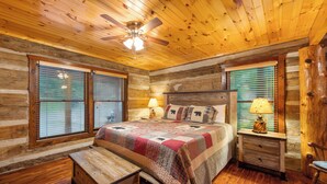 Un dormitorio de cabaña de troncos con techo de madera, una cama con colcha, dos mesitas de noche con lámparas y un sillón mecededor.