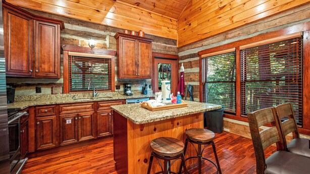 Cocina de cabaña de madera con gabinetes de madera, encimeras de granito, una isla central con taburetes y ventanas amplias con persianas.