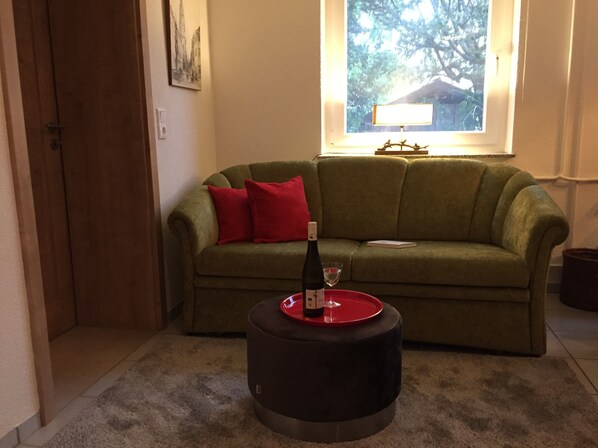 Un salon avec un canapé vert, des coussins rouges, un petit pouf rond, une bouteille de vin et un verre posé sur le pouf.