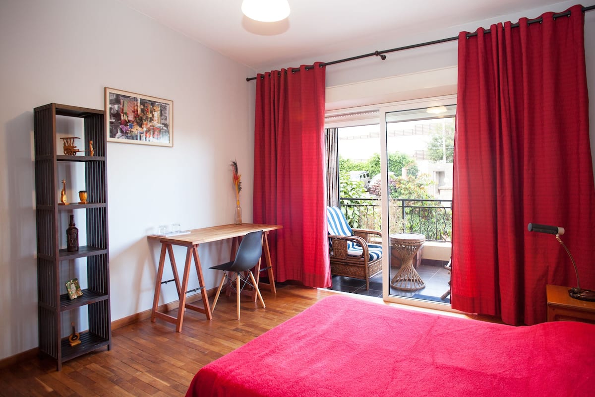 A bedroom with a bed, a desk, a chair, a bookshelf, and a balcony with red curtains.