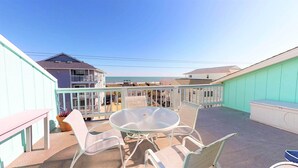 Une terrasse avec une table ronde en verre et quatre chaises, donnant sur une plage avec des bâtiments en arrière-plan.