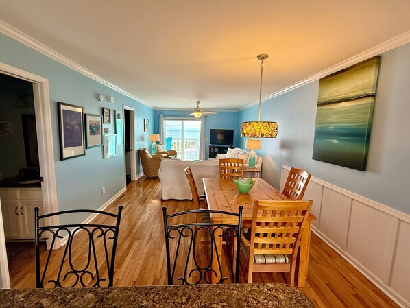 A dining area with a wooden table and chairs, a television in the background, and a view of the ocean through a sliding door.