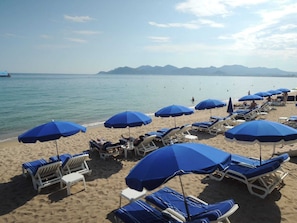 A beach with rows of sun loungers and blue umbrellas, a clear sky, and distant mountains.
