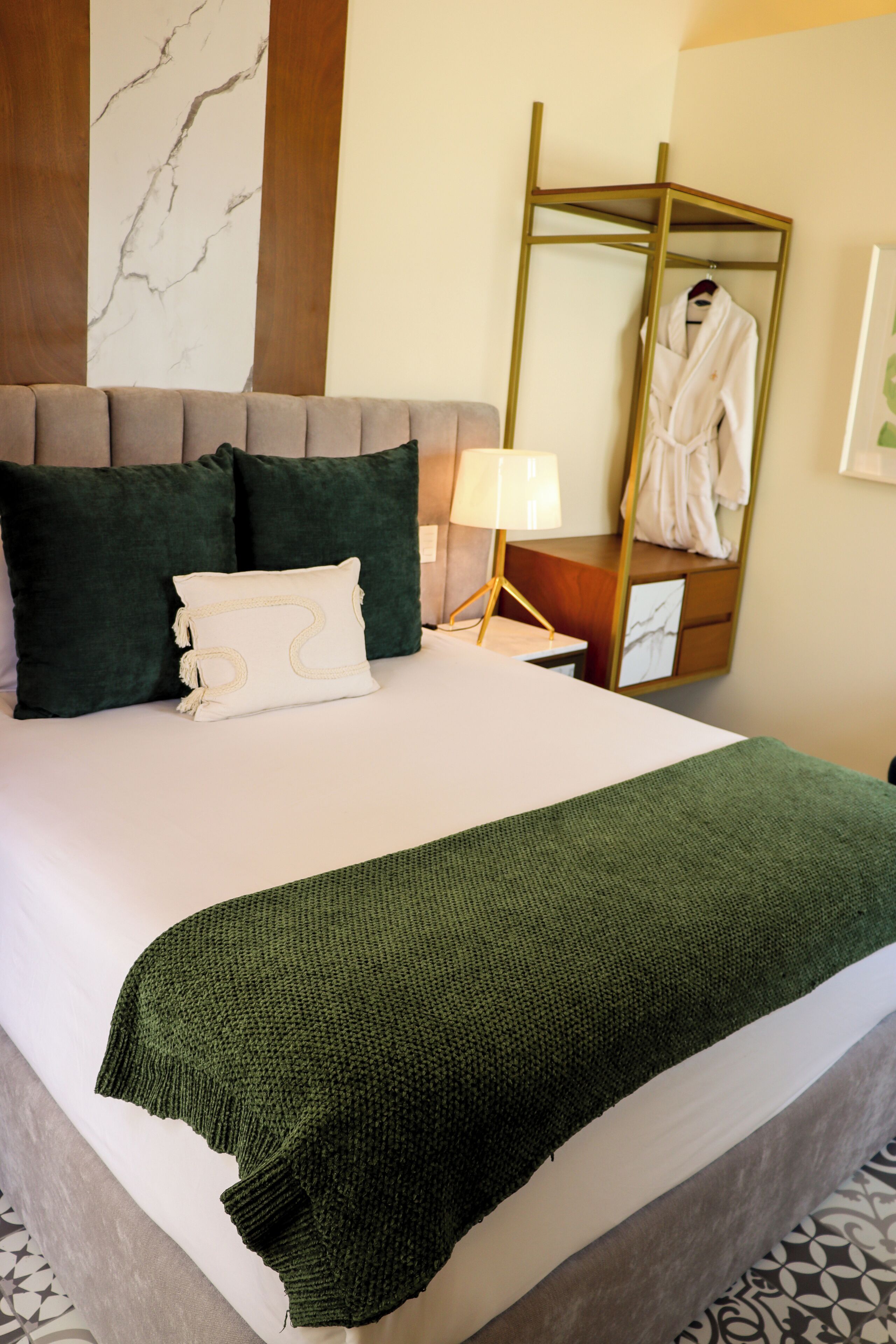 A neatly made bed with a green blanket, dark green pillows, and a white pillow with a design. There is a wooden headboard, a bedside table with a lamp, and a mirror on the wall.