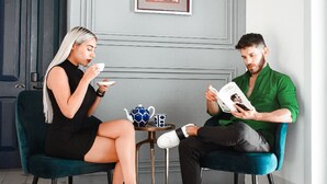 Deux personnes sont assises sur des chaises de couleur turquoise, autour d’une petite table sur laquelle se trouvent une théière et une tasse, dans une pièce dont la porte est grise et où un tableau encadré est accroché au mur.
