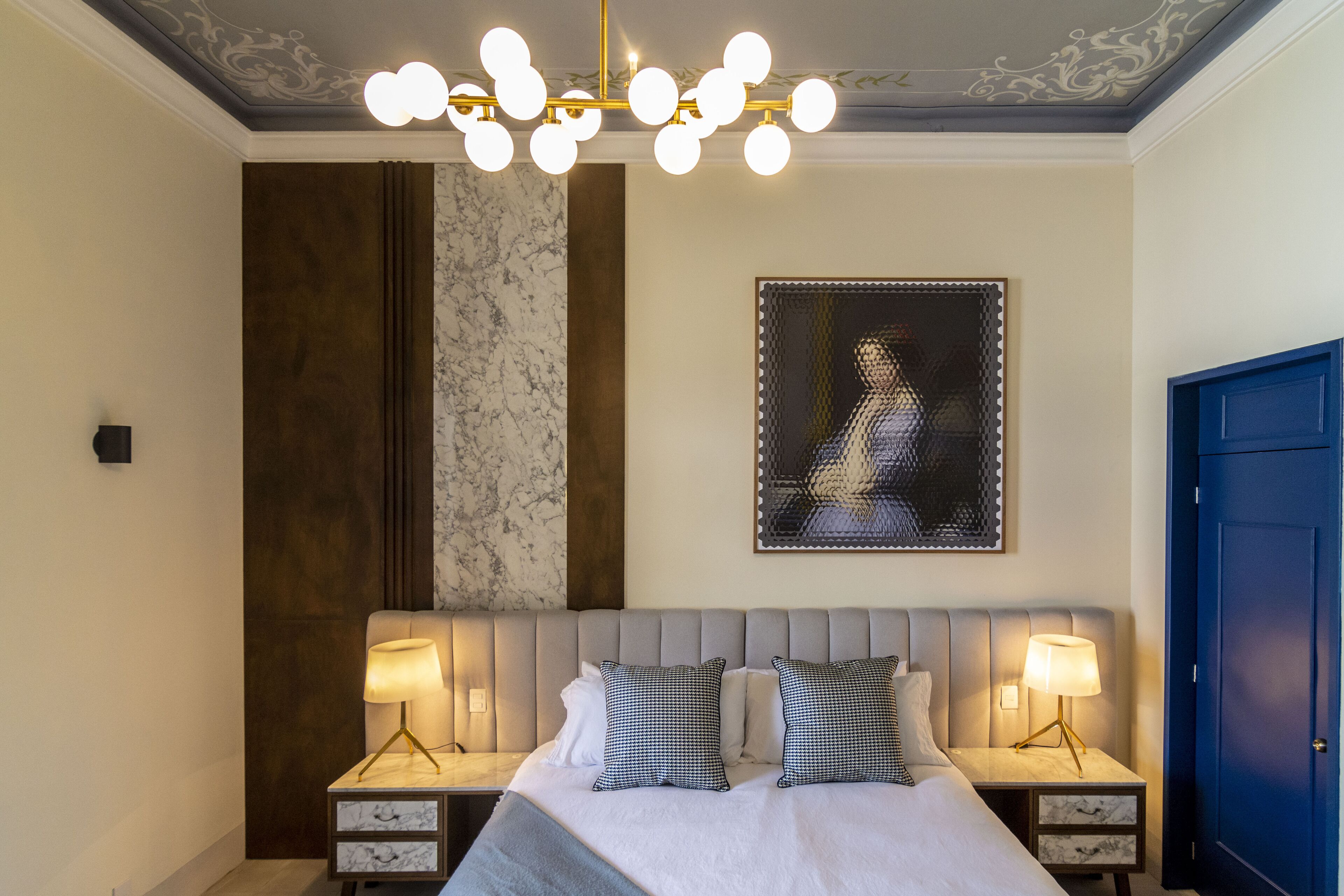 Una habitación de hotel con una cama grande, dos mesitas de noche con lámparas, una lámpara decorativa en el techo, una obra de arte enmarcada en la pared y una puerta azul.