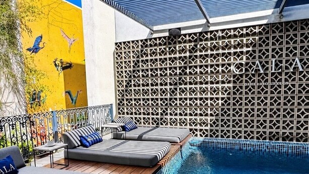 Une piscine avec un sol carrelé, un espace détente couvert avec des coussins et un mur décoré de motifs.
