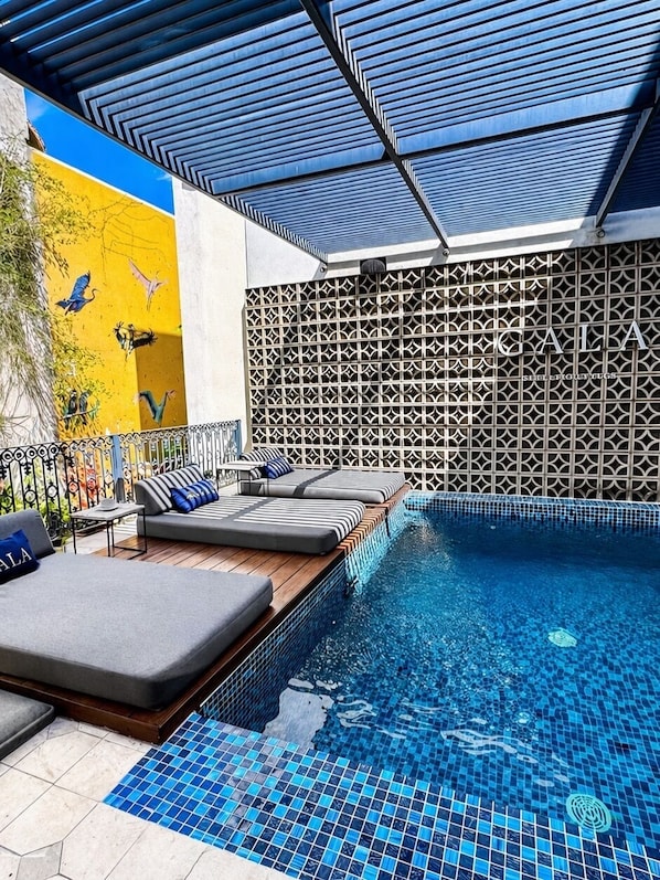 A swimming pool with a tiled floor, a covered seating area with cushions, and a patterned wall.