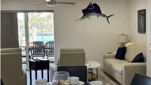 Ein Essbereich mit einem Glastisch, Stühlen und weißen Schüsseln. Es gibt einen Deckenventilator, eine Couch mit Kissen und einen Blick durch eine Glastür.