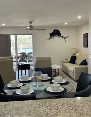 Un coin repas avec une table en verre, des chaises et des bols blancs. On y trouve également un ventilateur de plafond, un canapé avec des coussins et une vue à travers une porte vitrée.