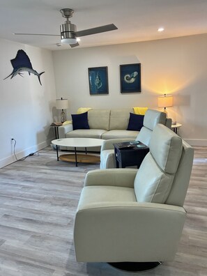 A modern living room with a beige sofa, a small round coffee table, and a ceiling fan. There are framed pictures on the wall and a lamp on a side table.