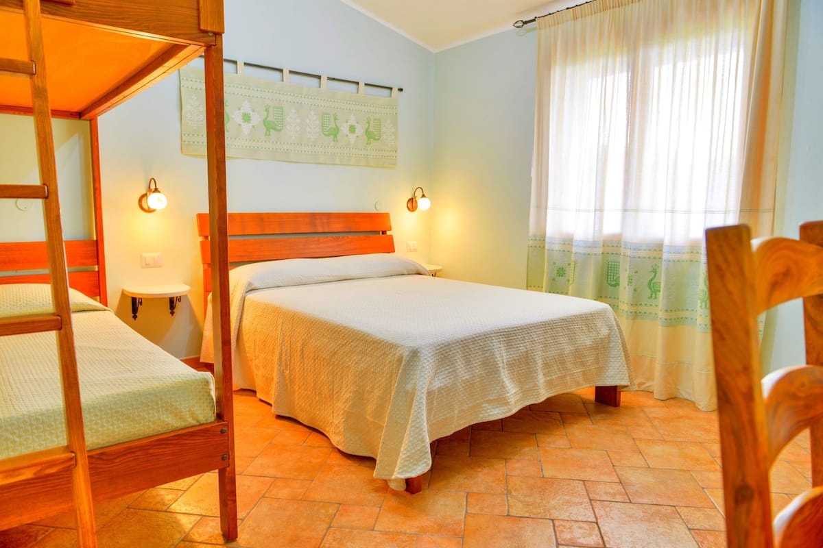 Un dormitorio con una cama litera de madera, una cama individual, dos lámparas de noche, una mesita de noche pequeña y una ventana con cortinas.