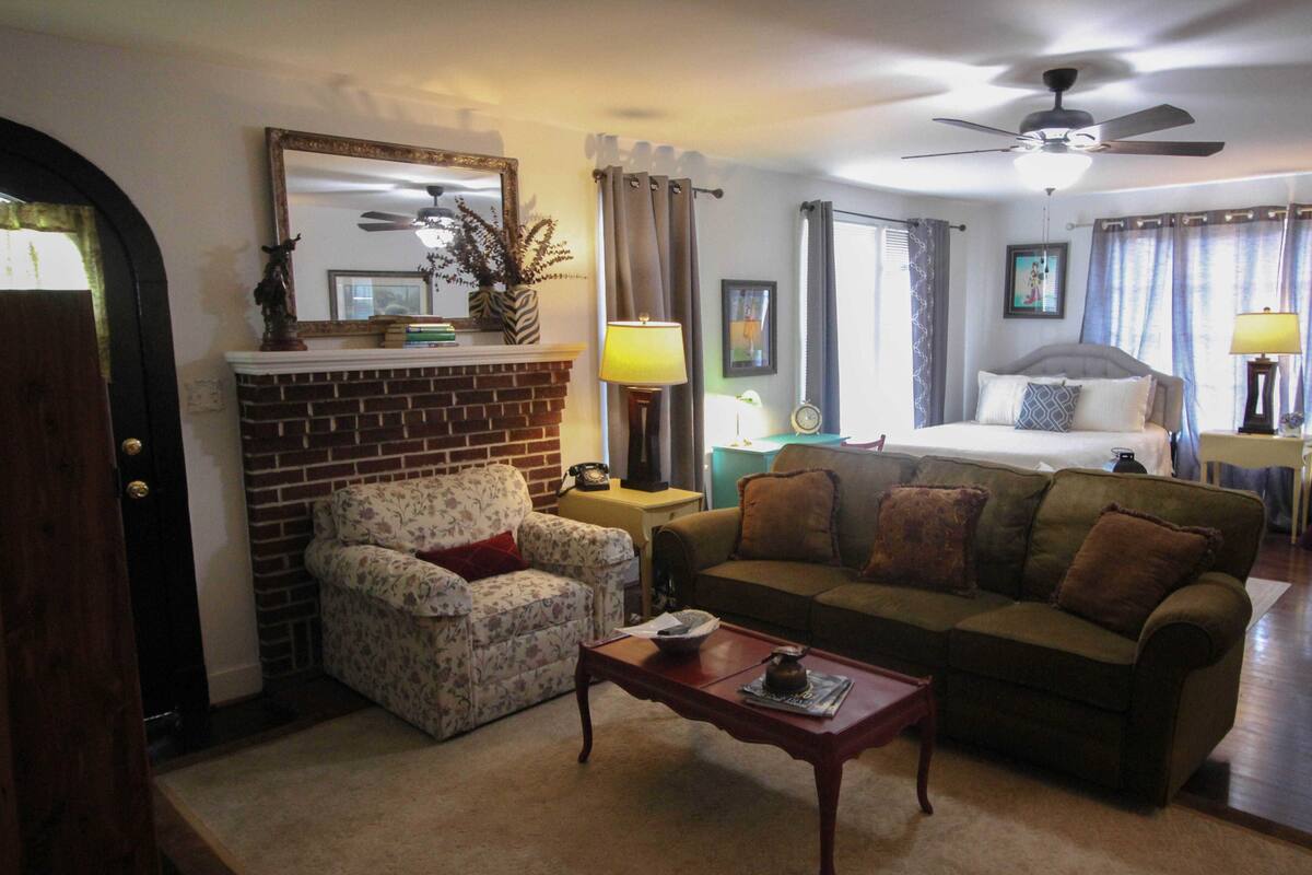 A hotel room with a brick fireplace, floral armchair, sofa, and a bed with a headboard.