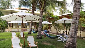 Un espacio al aire libre tropical con sillones blancos, sombrillas, palmeras y una hamaca.