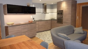 Una cocina compacta con electrodomésticos modernos, una zona de comedor con mesa de madera y un sofá gris.