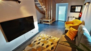 Una sala de estar con una televisión de pantalla plana, una escalera, un sillón amarillo y un sofá gris.