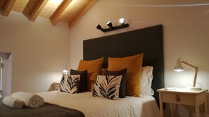 A neatly made bed with a zebra-patterned pillow, a white pillow, and a brown pillow. There is a bedside table with a lamp and a small drawer. The room has wooden beams on the ceiling and a dark-colored headboard. - Villa overlooking the countryside and castle (Óbidos)