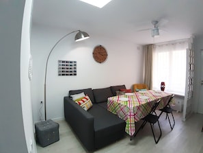 Un espace de vie compact comprenant un canapé, une table à manger avec des chaises, un ventilateur de plafond, une horloge murale et une fenêtre avec des rideaux.