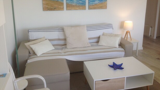 A cozy living room with a sofa, a coffee table, and a lamp. There are three paintings on the wall depicting a beach scene. - Apartment with balcony and terrace near the center and the beach (Royan)