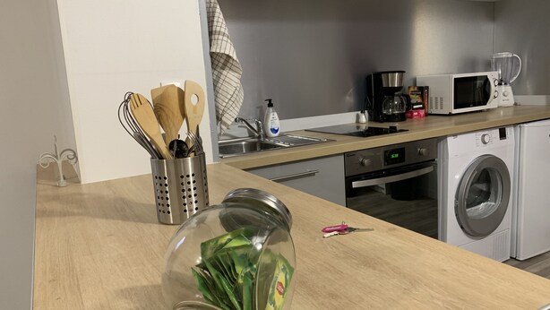 Een keuken met een magnetron, koffiezetapparaat en wasmachine. Op het aanrecht staat een glazen pot met groene bladeren.