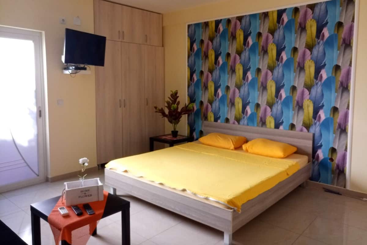 A bedroom with a bed, a television on the wall, a small table with a vase, and a colorful wall decoration.
