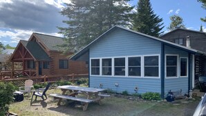 A rustic cabin with a picnic table and chairs, surrounded by a wooden deck and a small garden. - Gebhardt Getaway - Charming LAKEFRONT Cottage on Lake Willoughby (Westmore)
