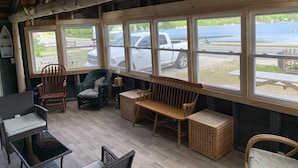 A cozy room with a wooden ceiling, large windows overlooking a body of water, and a seating area with wicker furniture and a wooden bench. - Gebhardt Getaway - Charming LAKEFRONT Cottage on Lake Willoughby (Westmore)