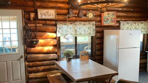 A rustic wooden cabin interior with a dining table, benches, a refrigerator, and a cozy seating area. - Gebhardt Getaway - Charming LAKEFRONT Cottage on Lake Willoughby (Westmore)