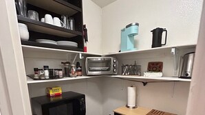 A compact kitchen with a microwave, toaster, and coffee maker on shelves, a small refrigerator, and a wooden cutting board on the counter.