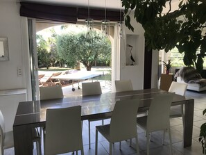 Ein moderner Essbereich mit einem Glastisch und weißen Stühlen, ein großes Fenster mit Blick auf einen Garten und einer deckenmontierten Leuchte.