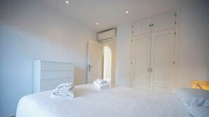 Un lit bien fait, avec des serviettes pliées, une armoire blanche et un appareil de climatisation placé au-dessus d’une porte.