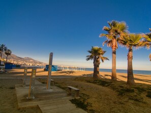 A beach with palm trees, a wooden boardwalk, and a distant cityscape.