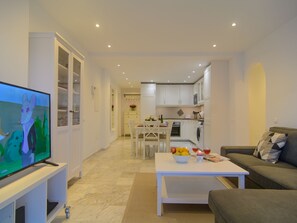 Una sala de estar moderna con una televisión de pantalla plana, un sofá, una mesa de centro con frutas y bebidas, y una cocina al fondo.