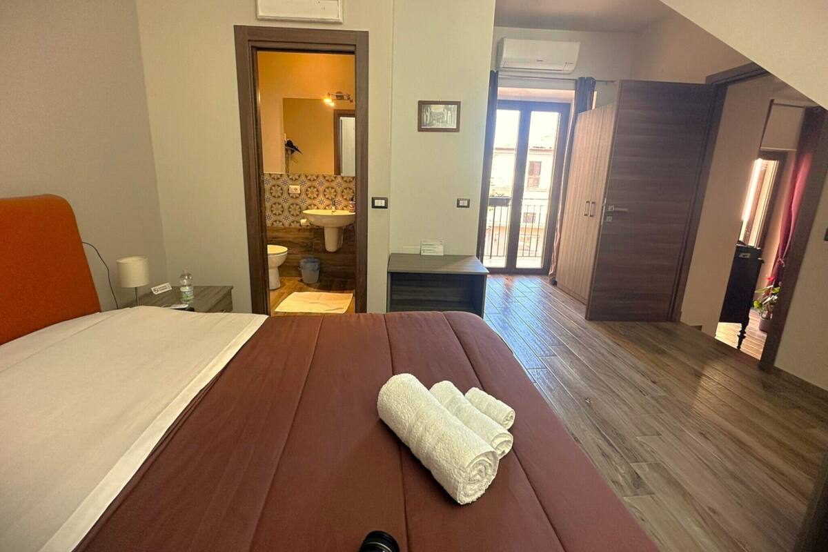 A hotel room with a bed, a wooden floor, a bathroom visible through an open door, and a view of the outside balcony.