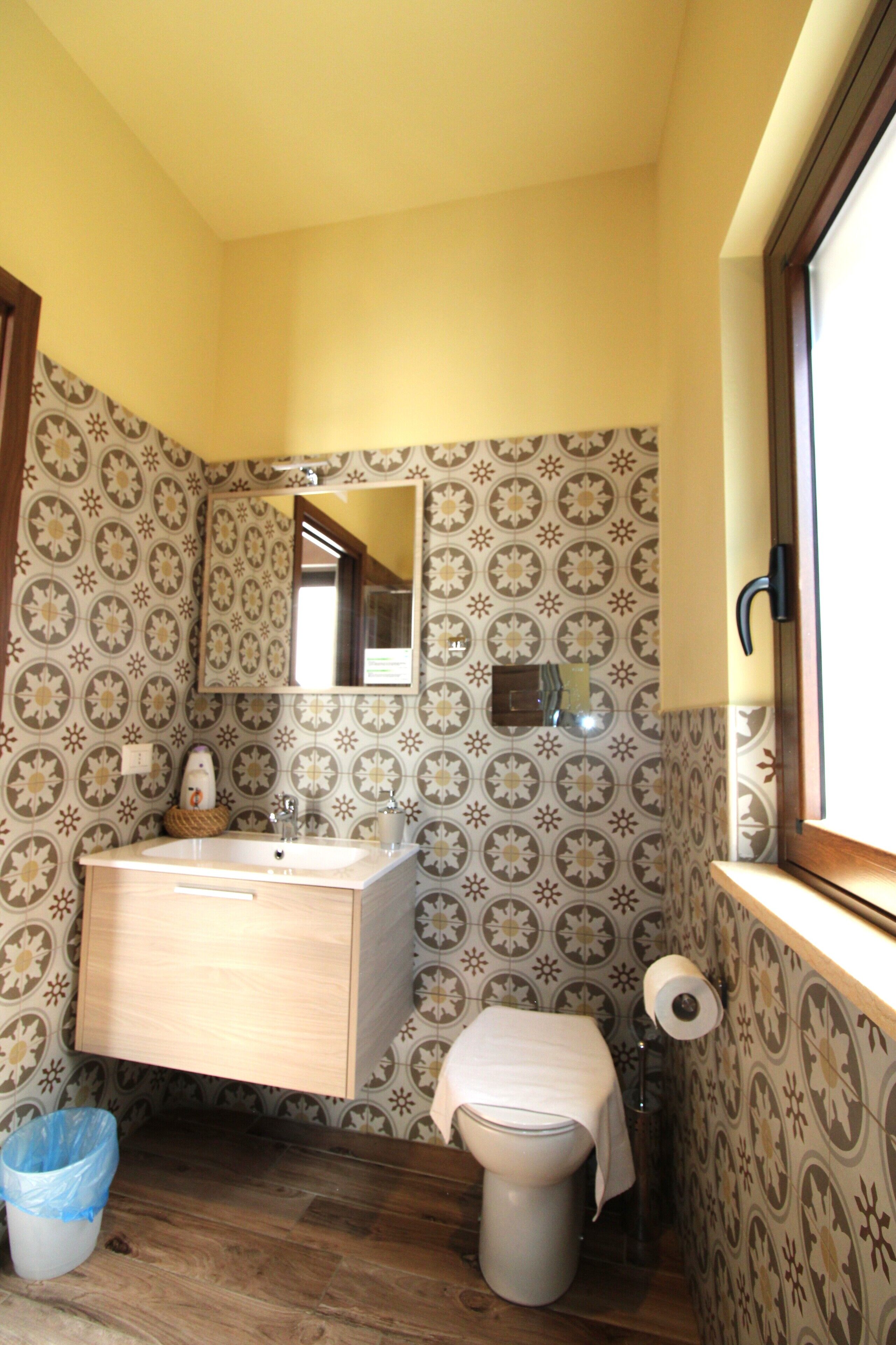 Casa de banho com papel de parede estampado, lavatório de madeira, espelho, sanita e janela com cortina.