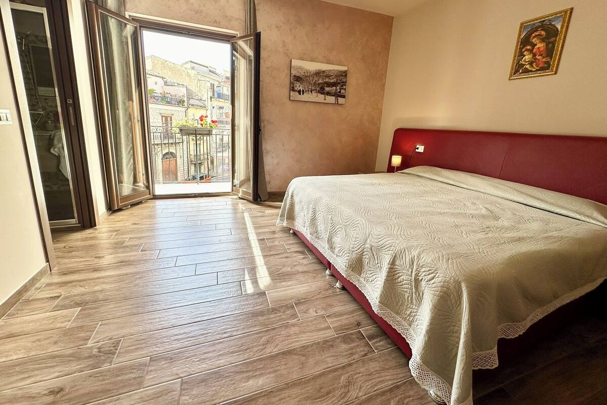 A hotel room with a bed, a balcony with a view of buildings, and a painting on the wall.