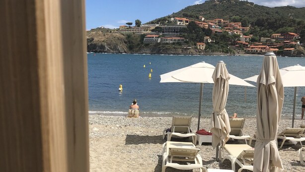 A beach with sun loungers and umbrellas, a clear sky, and buildings in the background. - Charming in Collioure – where creativity meets the beach (Collioure)