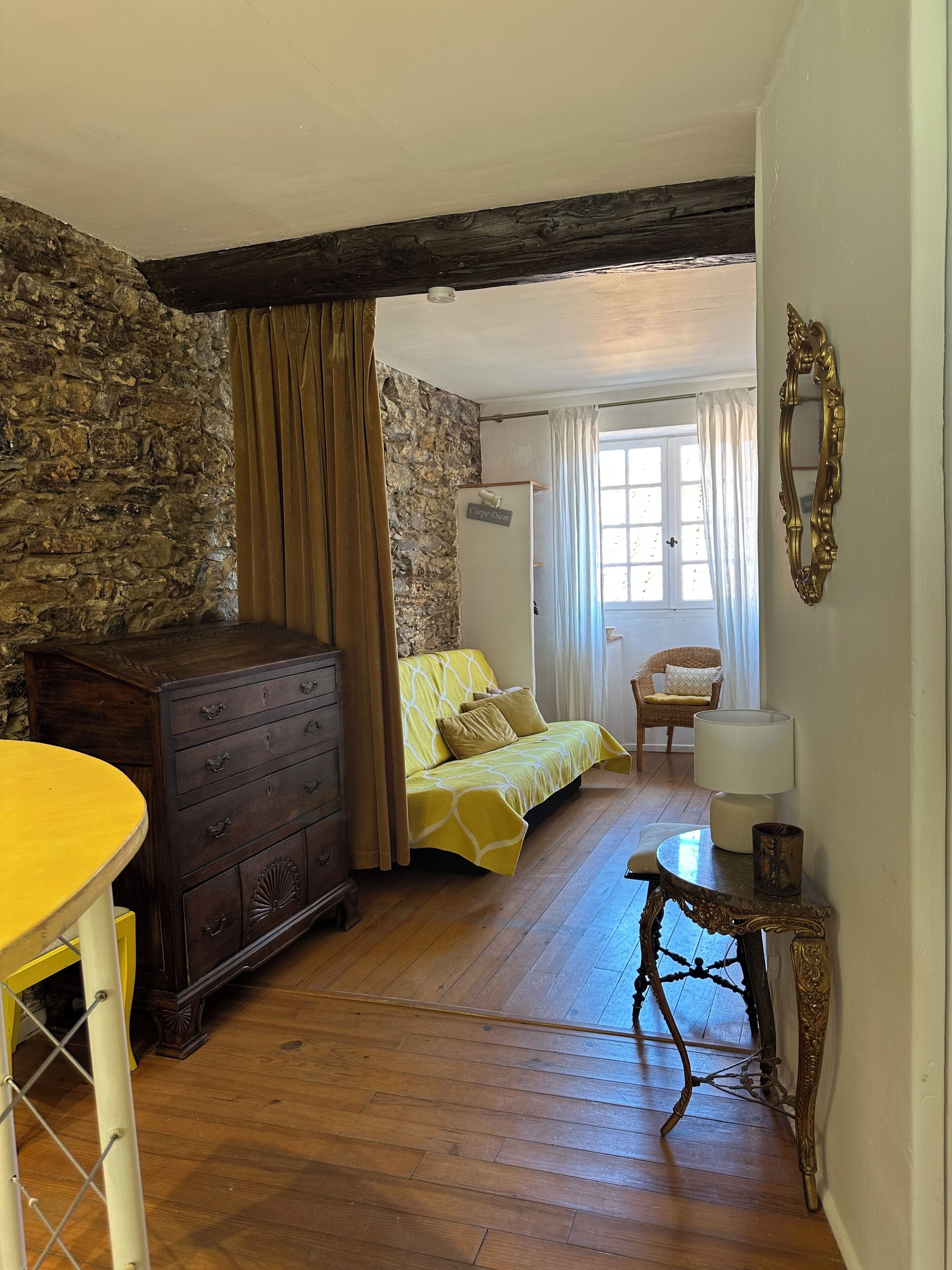 A room with a stone wall, wooden floor, a bed with yellow bedding, a wooden dresser, a table with a yellow top, and a mirror on the wall.