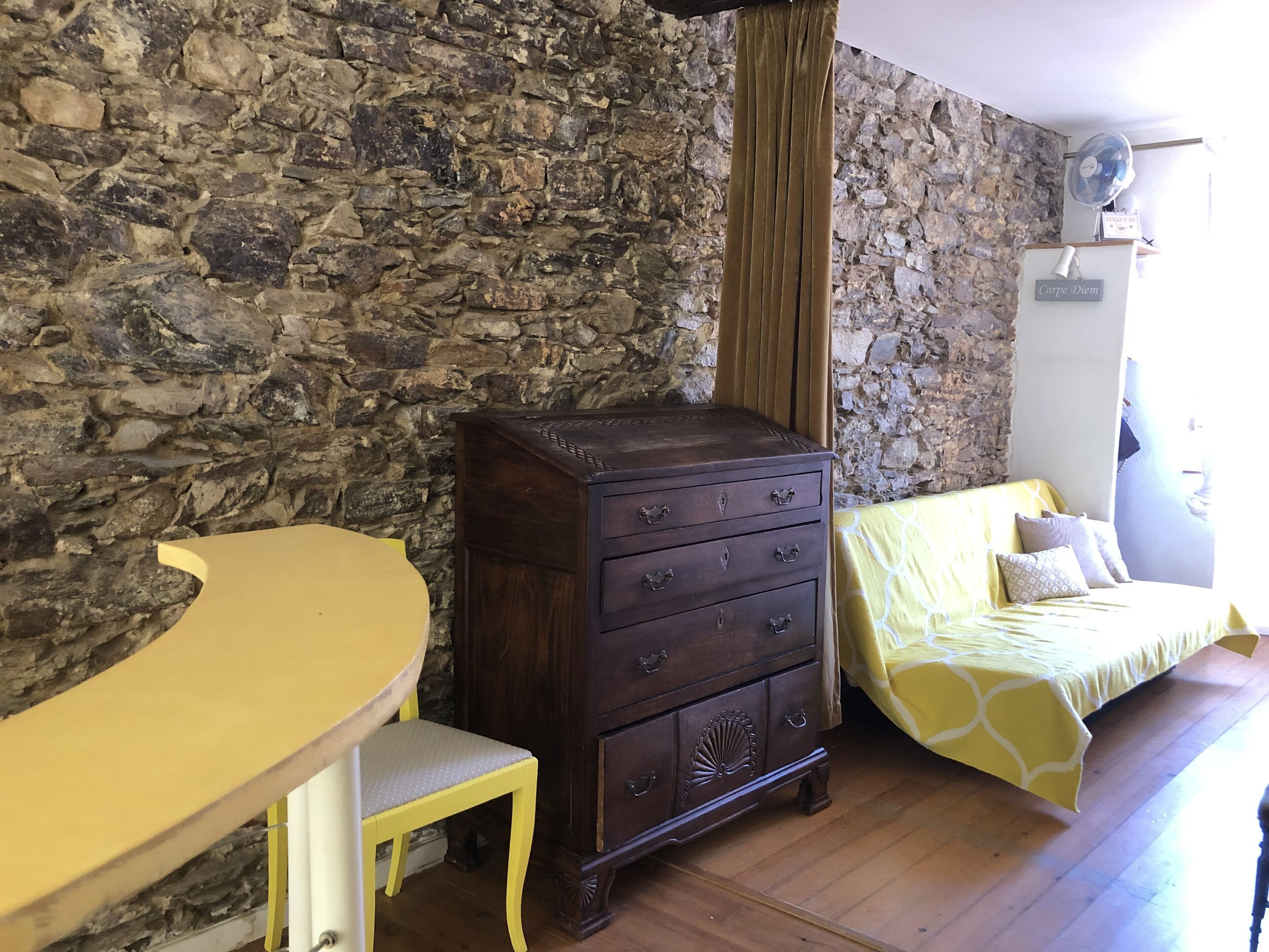 Una habitación con pared de piedra, cómoda de madera, una mesa amarilla y una cama con colcha de diseño amarillo y blanco.