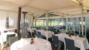 Un área de comedor con carpas, mesas y sillas redondas, ventanas amplias y un candelabro central.