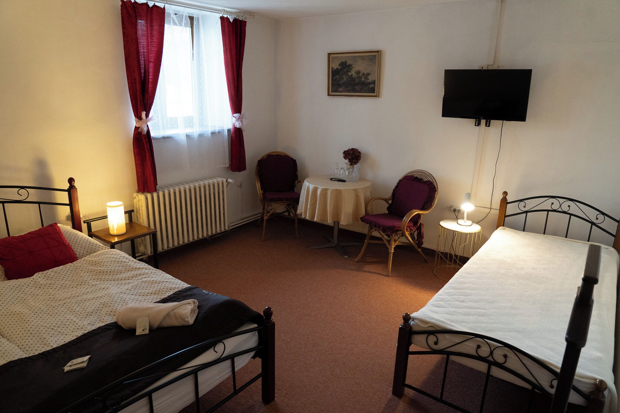 A room with two beds, a table with a vase, a chair, a television, and a radiator.