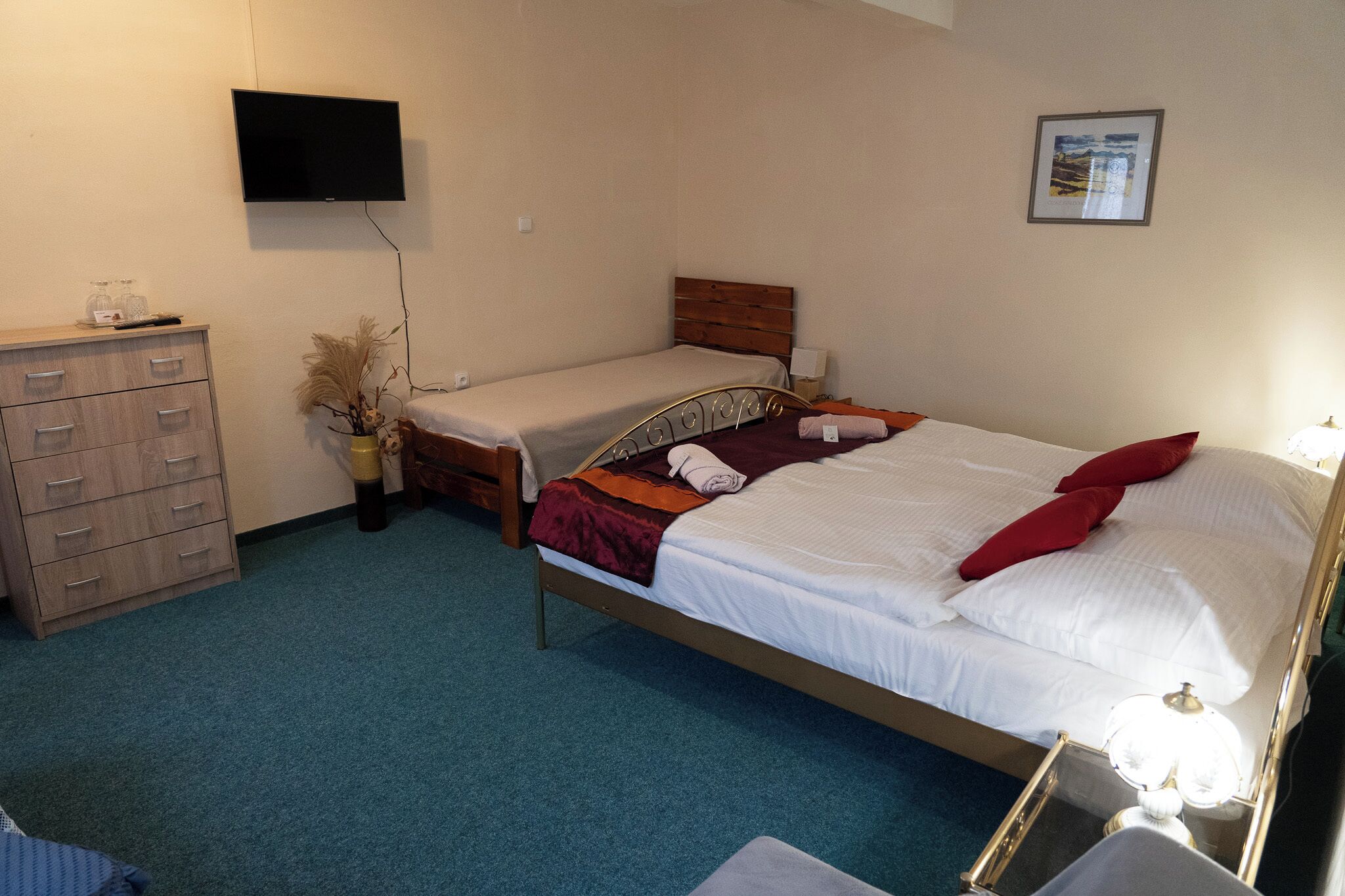 Une chambre avec un lit simple, une commode, un téléviseur fixé au mur et un tableau encadré.